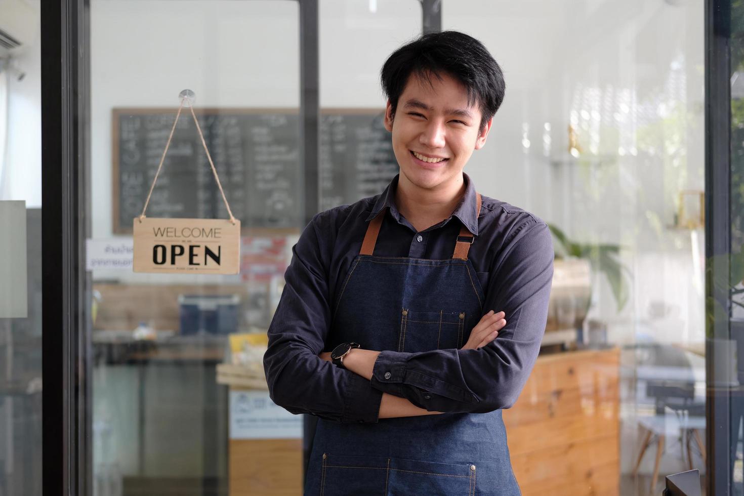 Happy asian young barista male in apron holding tablet and standing in front of the door of cafe with open sign board. Business owner startup SME entrepreneur concept photo