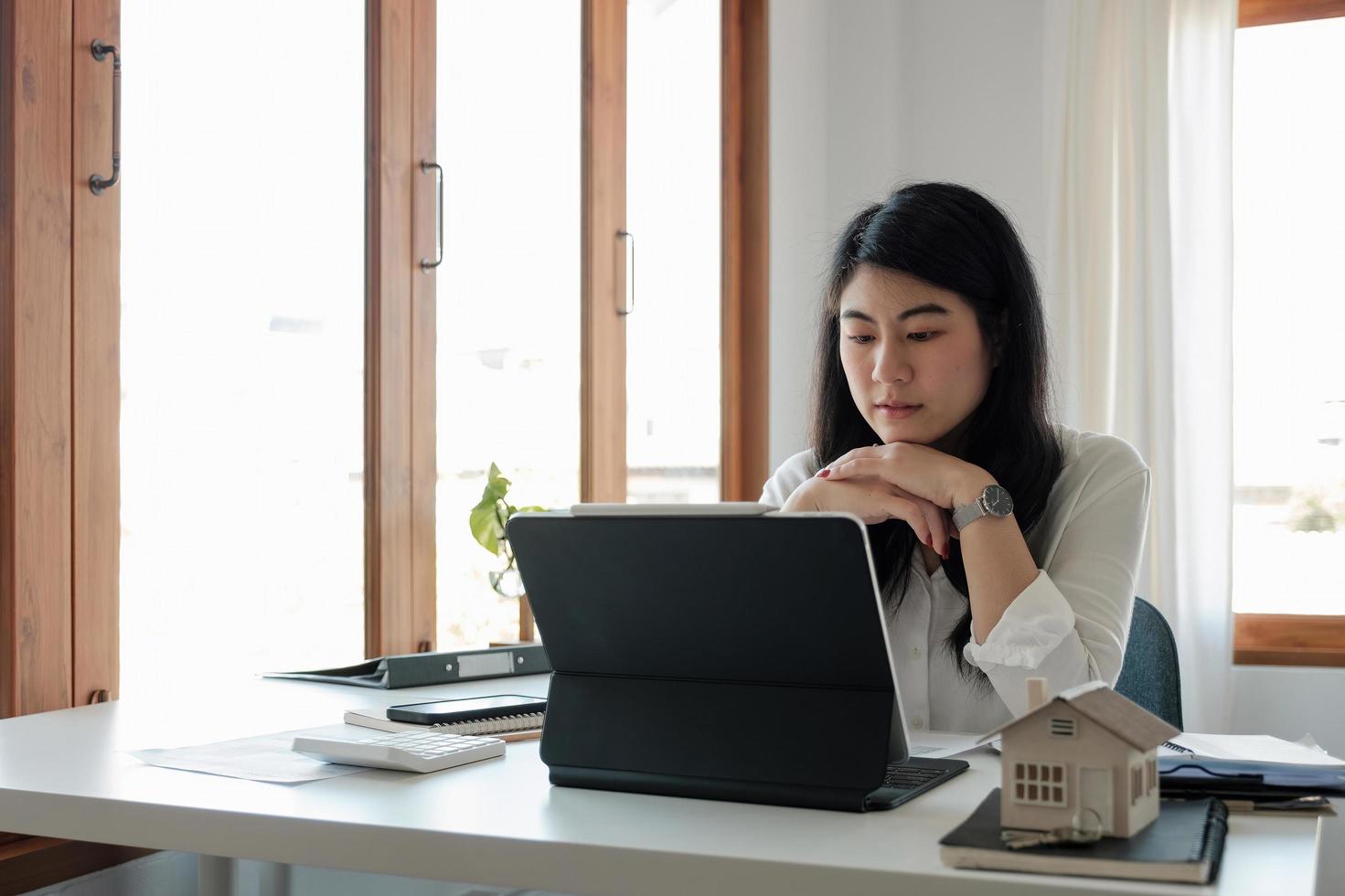 Close up confident asian woman looking at laptop screen, thoughtful student or businesswoman touching chin, pondering idea, strategy, working on research project in kitchen at home photo