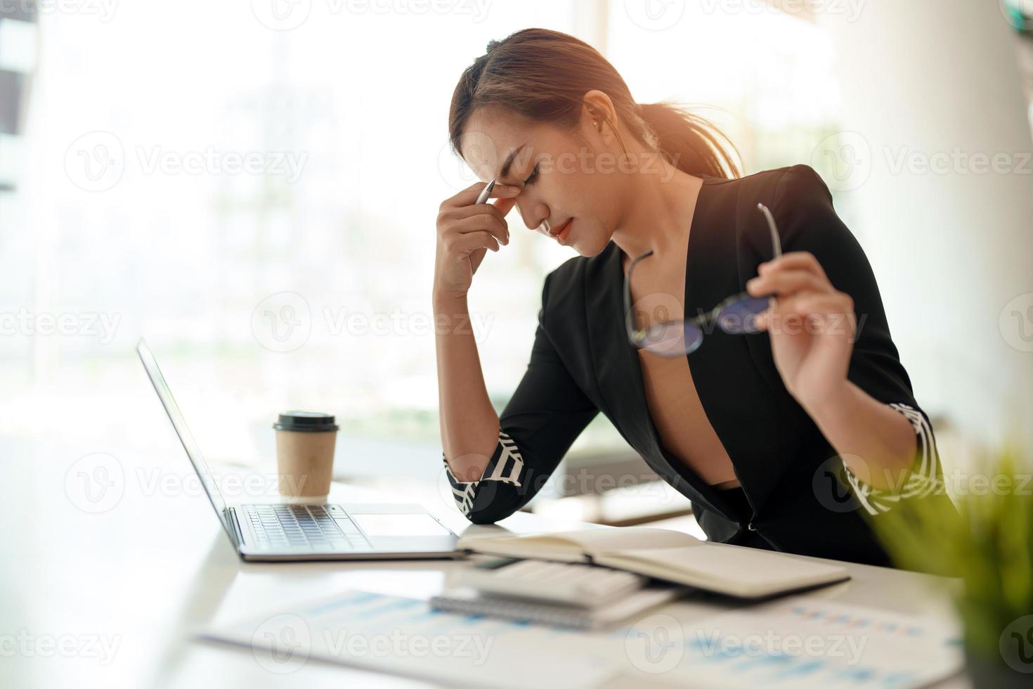 mujer asiática de negocios muy estresada sentada frente a su computadora mirando una gran pila de papeleo, mientras sostenía una mano en su frente foto