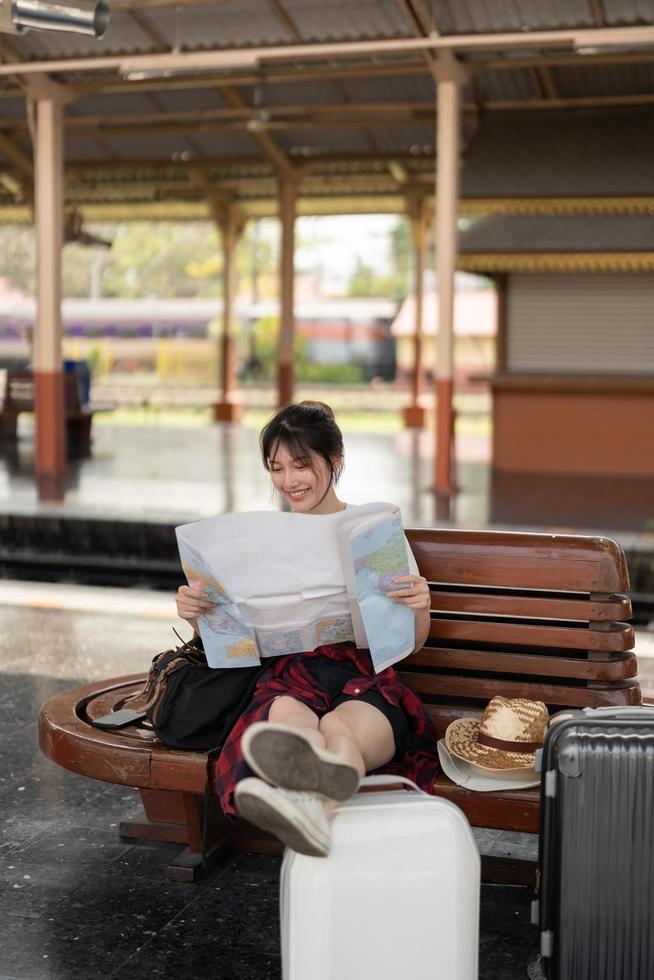 Happy young asian woman traveler or backpacker using map choose where to travel with luggage at train station, summer vacation travel concept photo