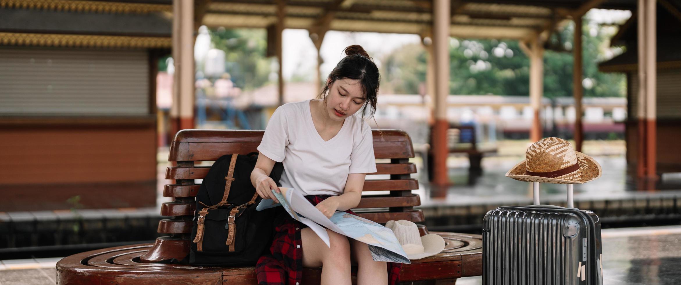 young asian woman traveler sitting with map choose where to travel and bag waiting for train at train station, summer vacation travel concept photo