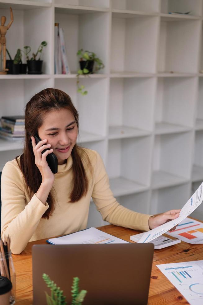 mujer de negocios asiática que hace una llamada telefónica, contadora que trabaja en inversiones financieras en calculadora con cálculo, análisis de negocios y crecimiento del mercado en gráficos y escritura de datos de documentos financieros. foto