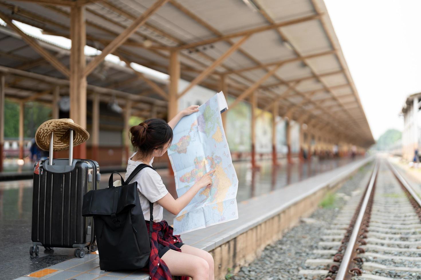 joven asiática usando un mapa local genérico, sentada sola en la plataforma de la estación de tren con equipaje. viaje de vacaciones de verano o joven turista mochila concepto de viajero foto