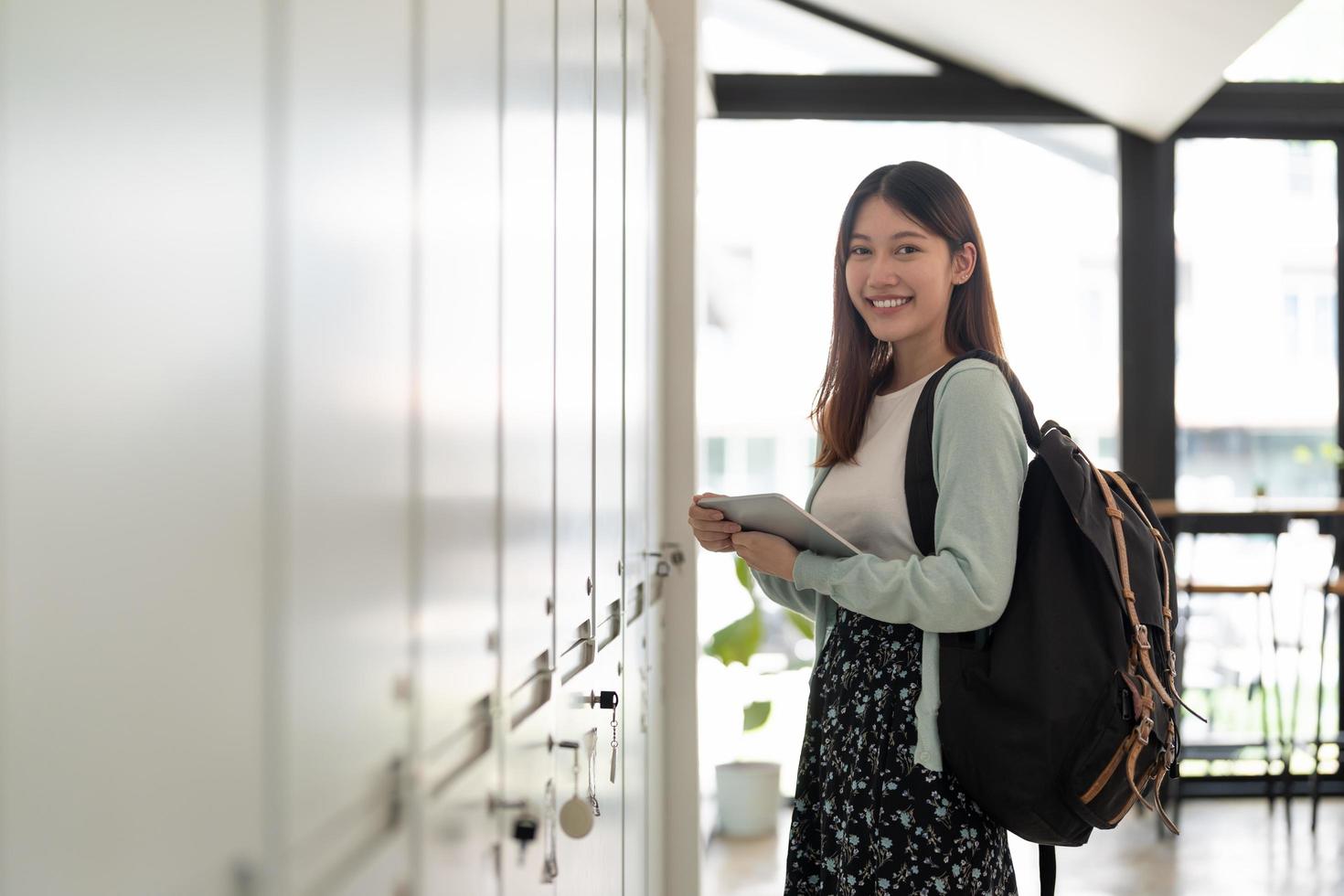 Portrait young asian woman student holding digital tablet at college standing in front of locker with backpack. photo