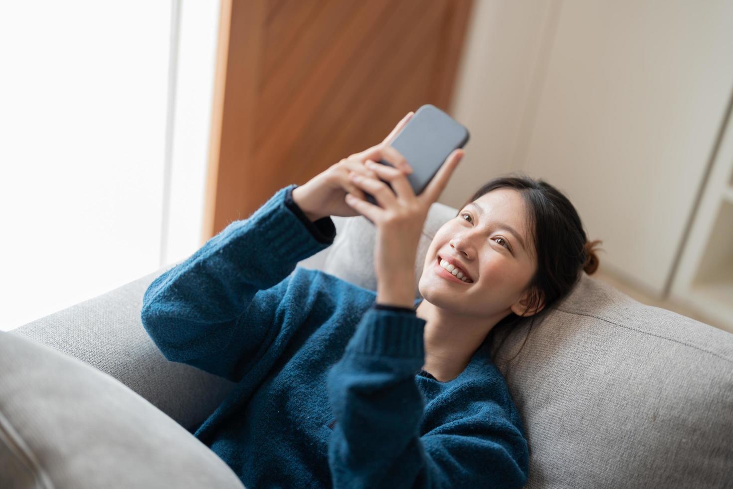 imagen de una atractiva mujer asiática sonriente usando un teléfono móvil mientras está acostada en un sofá en casa foto