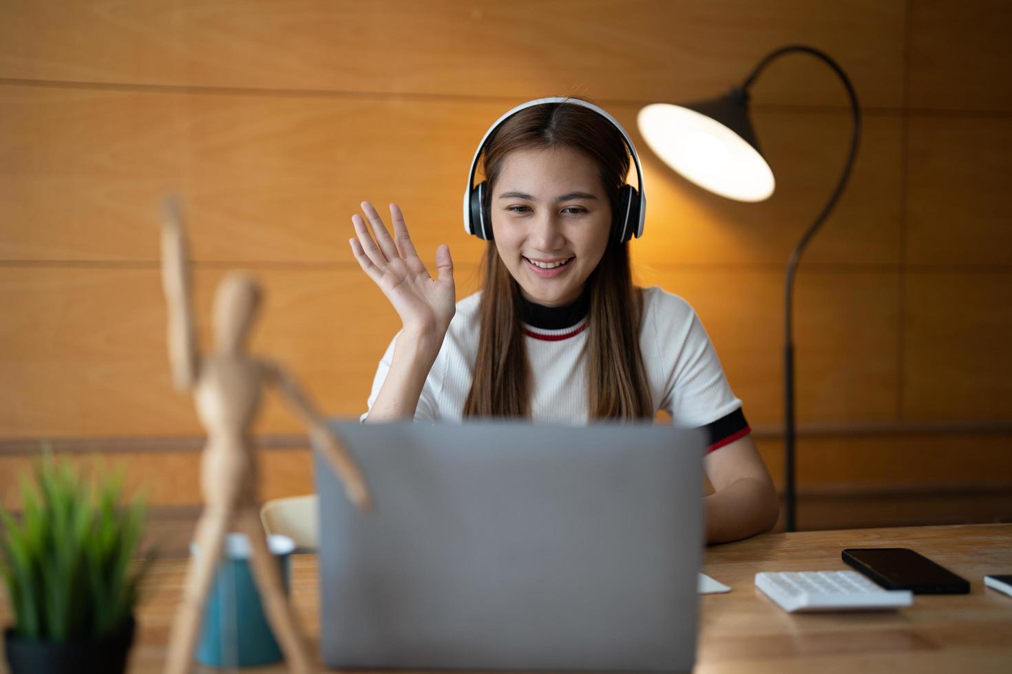 Smiling young asian woman in headset wave greet talking on webcam virtual conversation on laptop, happy female in wireless headphones speak on video call on computer, consult client online photo