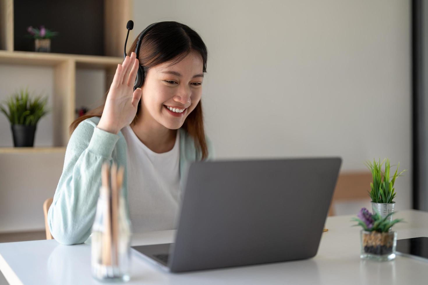 Attractive happy young student studying online at home, using laptop computer, headphones, having video chat, waving. Remote work, distance education. Video conference or virtual event on quarantine photo