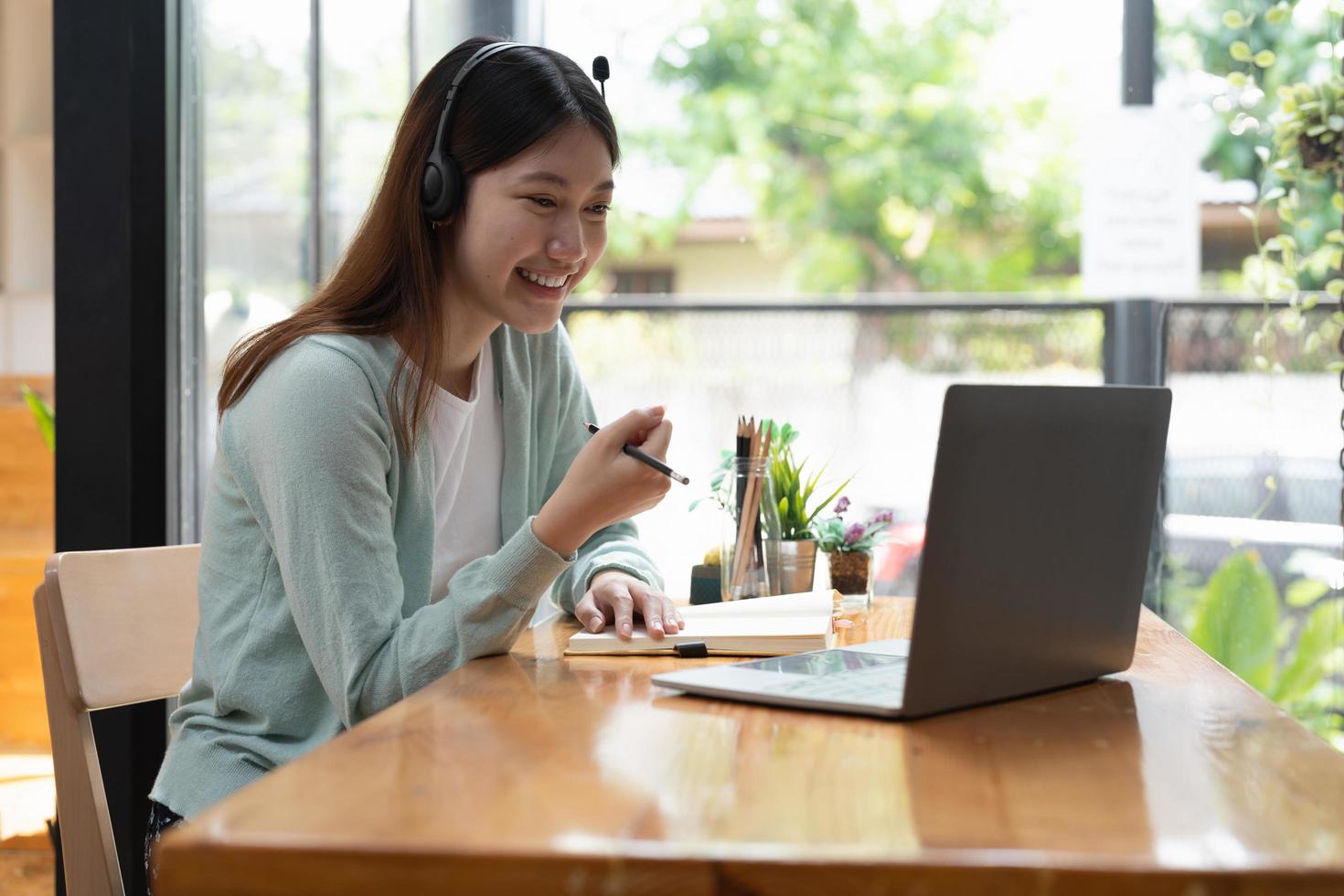 Smiling girl student wear wireless headphone study online with teacher, happy young asian woman learn language listen lecture watch webinar write notes look at laptop sit in cafe, distant education photo