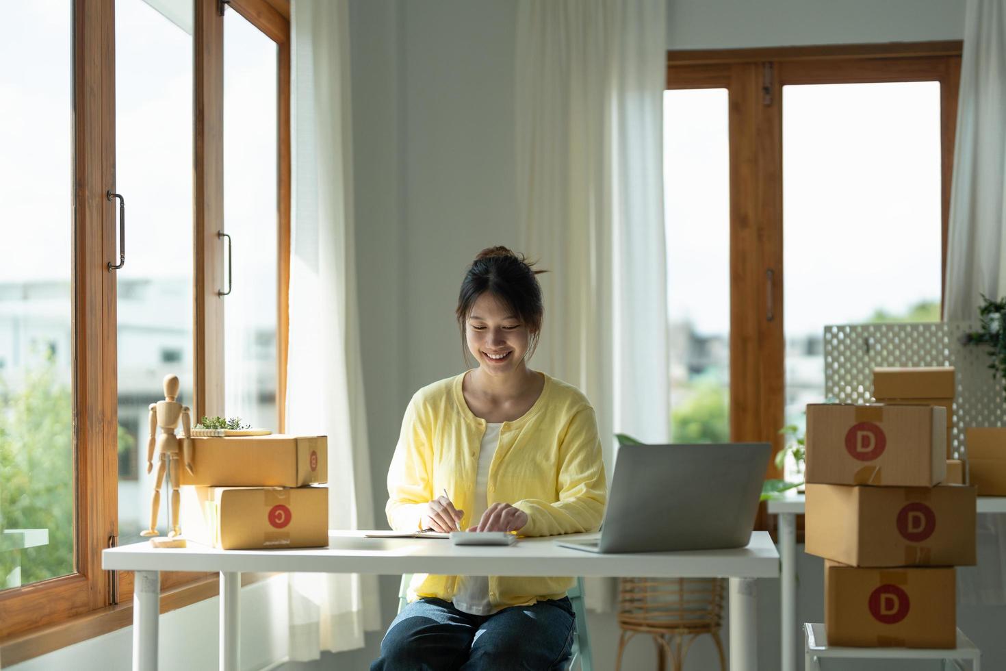 Close up asian female entrepreneur, checks online orders and uses a price calculator to calculate product prices and packs products into boxes to customers. photo