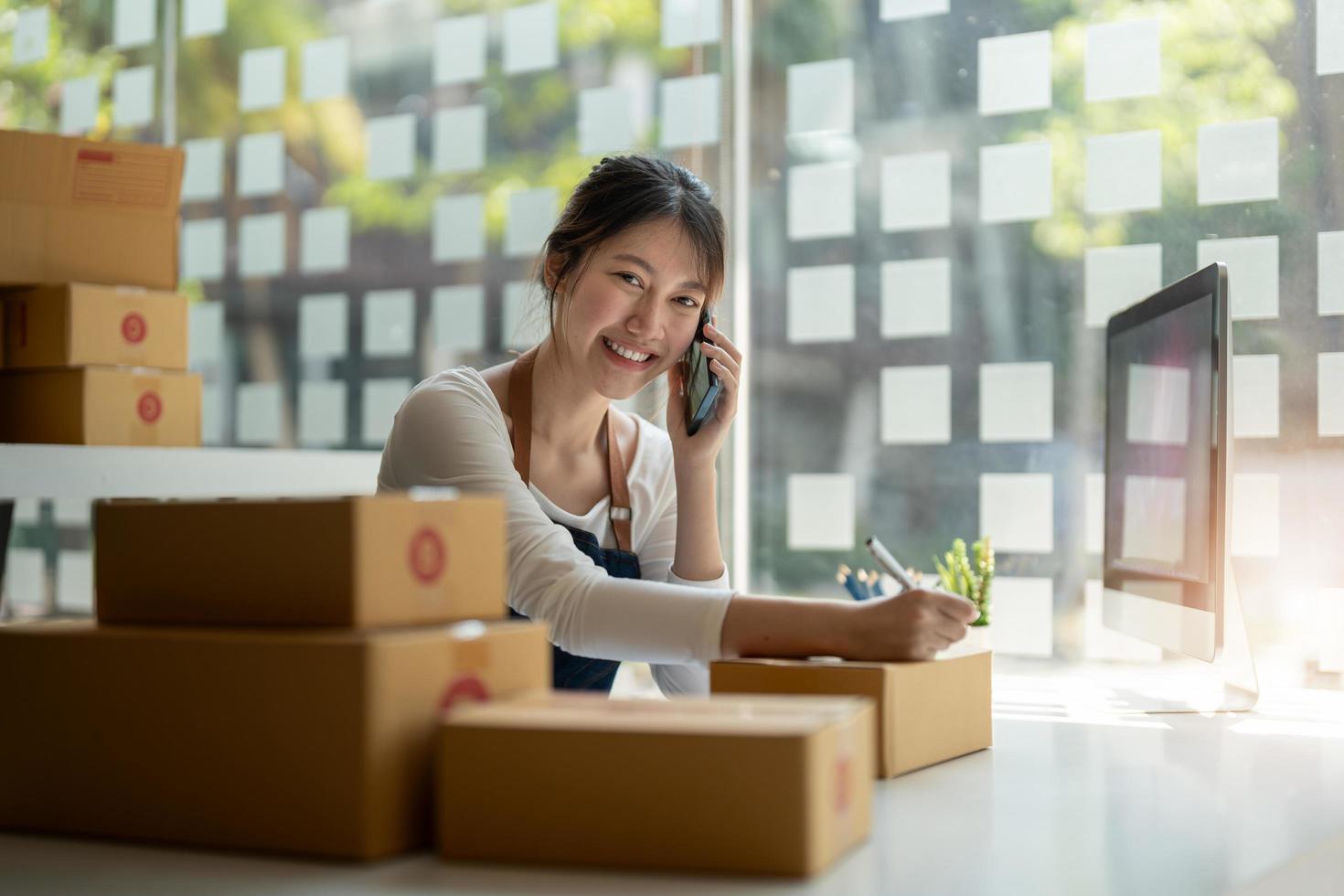 Young Asian small business owner working at home office, taking note on purchase orders. Online marketing packaging delivery, startup SME entrepreneur or freelance woman concept photo