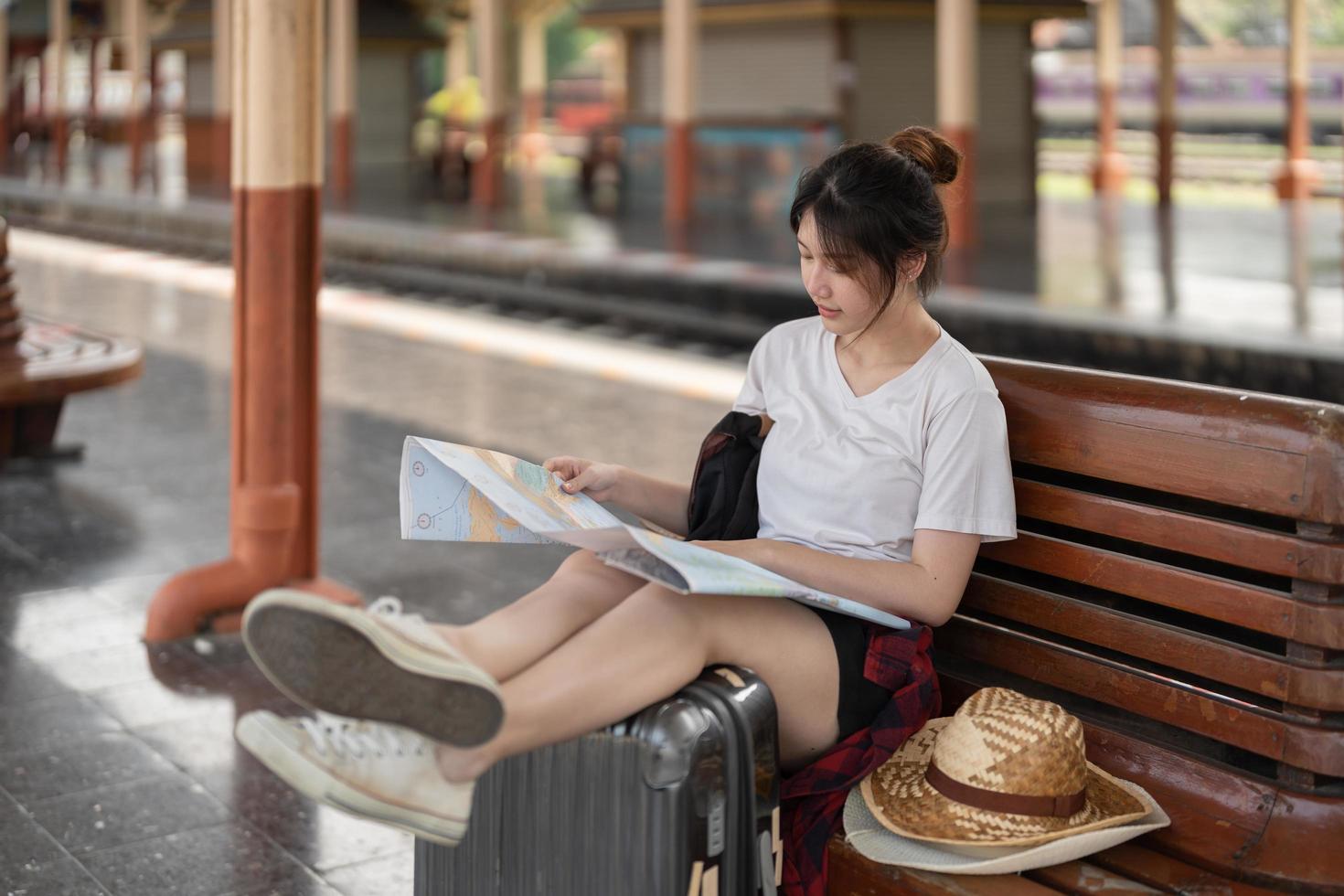 Happy young asian woman traveler or backpacker using map choose where to travel with luggage at train station, summer vacation travel concept photo