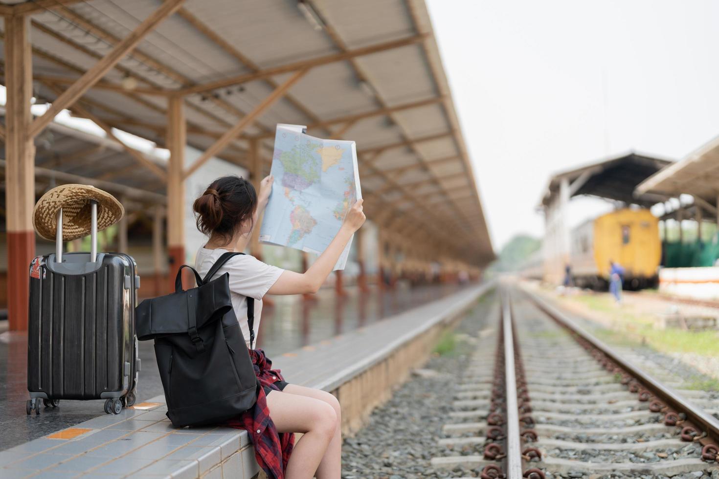 joven asiática usando un mapa local genérico, sentada sola en la plataforma de la estación de tren con equipaje. viaje de vacaciones de verano o joven turista mochila concepto de viajero foto
