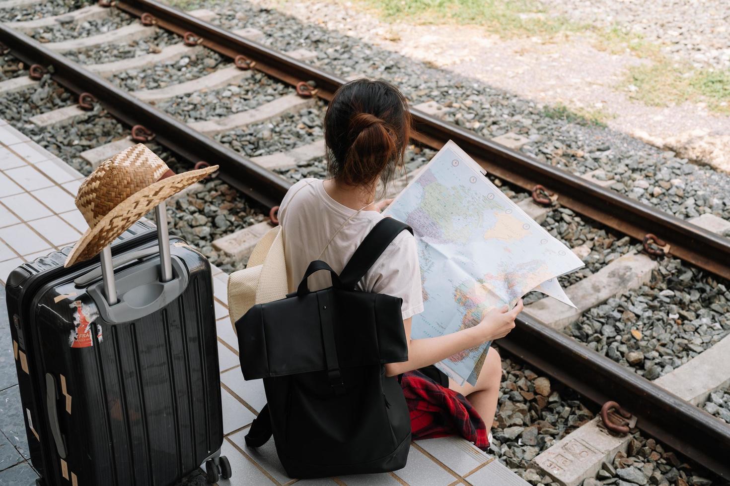 Young woman traveler with backpack looking to map while waiting for train, Asian backpacker on railway platform at train station. Holiday, journey, trip and summer summer travel concept photo