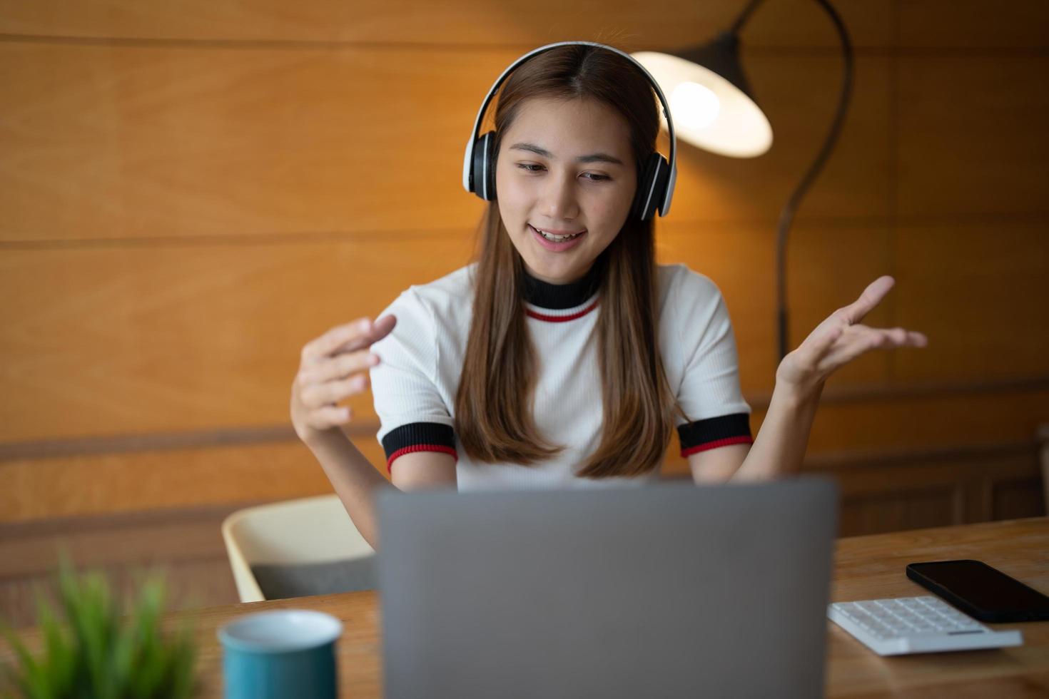 Smiling young asian woman in headset wave greet talking on webcam virtual conversation on laptop, happy female in wireless headphones speak on video call on computer, consult client online photo