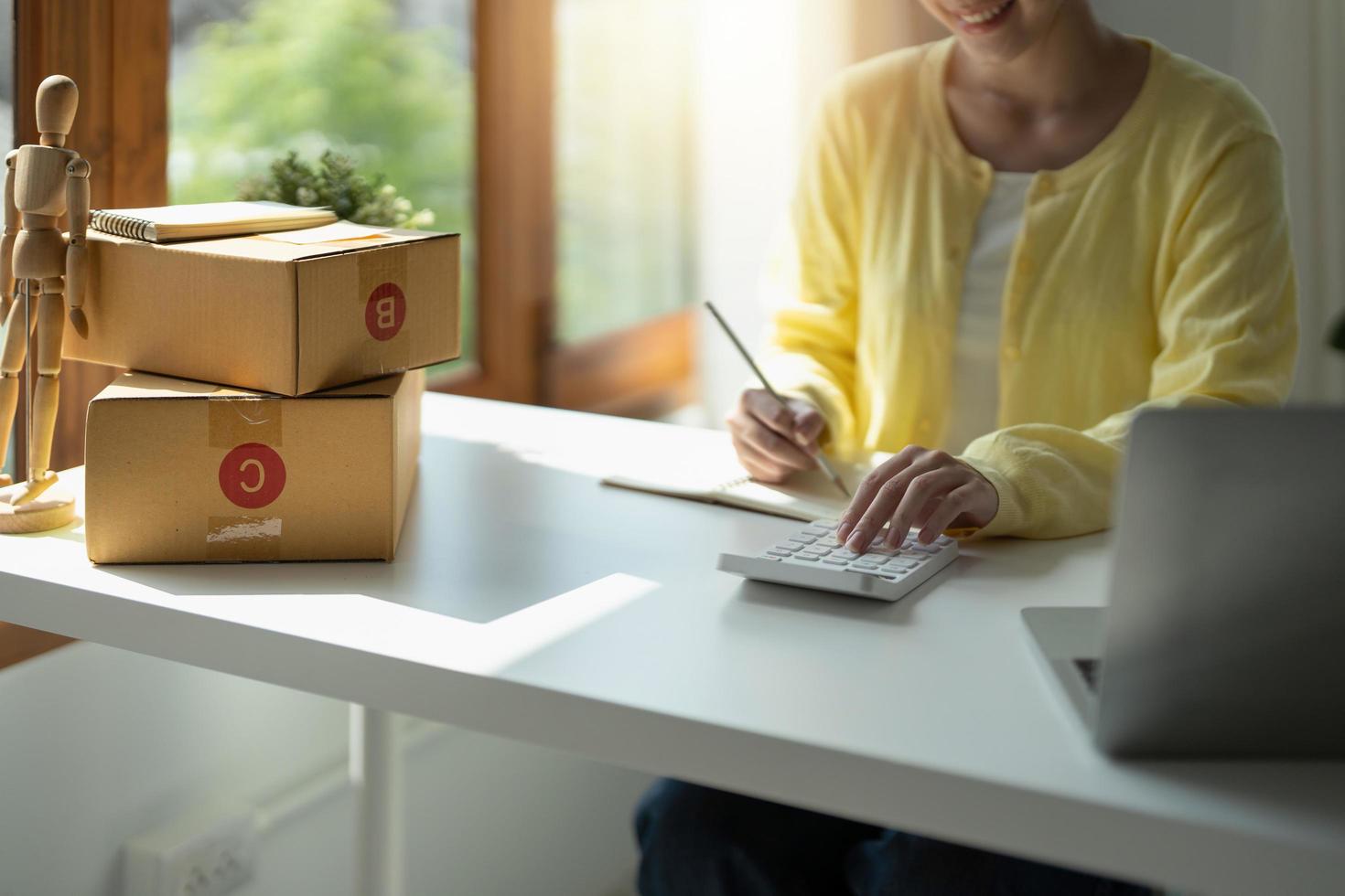 Close up asian female entrepreneur, checks online orders and uses a price calculator to calculate product prices and packs products into boxes to customers. photo