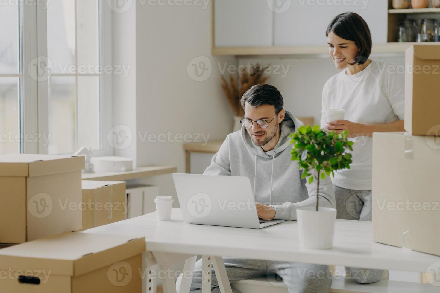 Indoor shot of happy woman and man spend morning at their new kitchen with big windows and modern furniture, husband works on laptop computer discusses with wife trendy design, surrounded with boxes photo