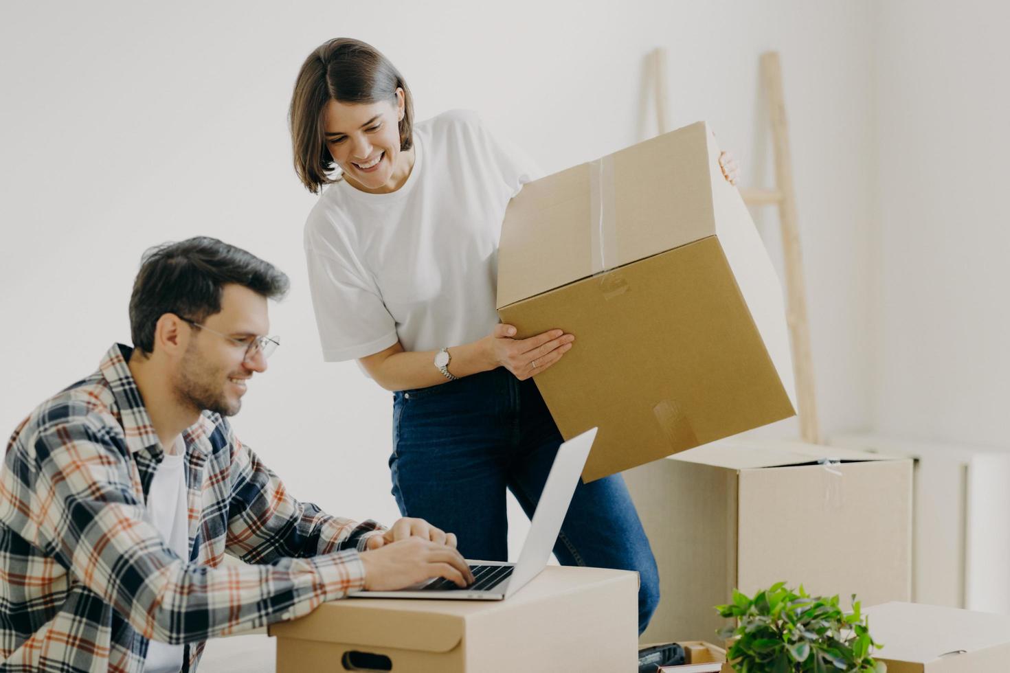 Freshly married family couple pose in their new apartment, unpack boxes with belongings, glad man sits in front of laptop computer, searches ideas for designing, have domestic routine problems photo