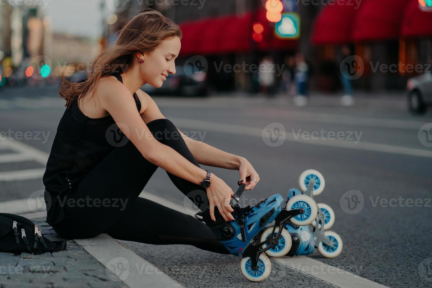 tiro al aire libre de mujer activa cordones patines se prepara para el paseo se sienta en la carretera contra el fondo de la ciudad ocupada vestida con ropa deportiva negra disfruta patinando. estilo de vida deportivo y concepto de recreación foto