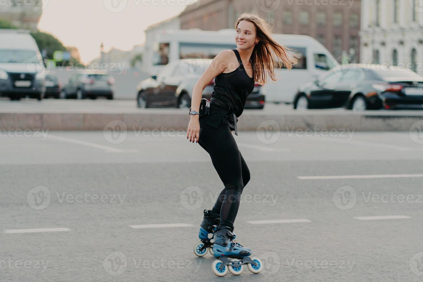 una foto lateral de una joven activa en patines en buena forma física a lo largo de las calles de la ciudad se ve feliz detrás, usa ropa deportiva negra y lleva un estilo de vida saludable. concepto de recreación y hobby