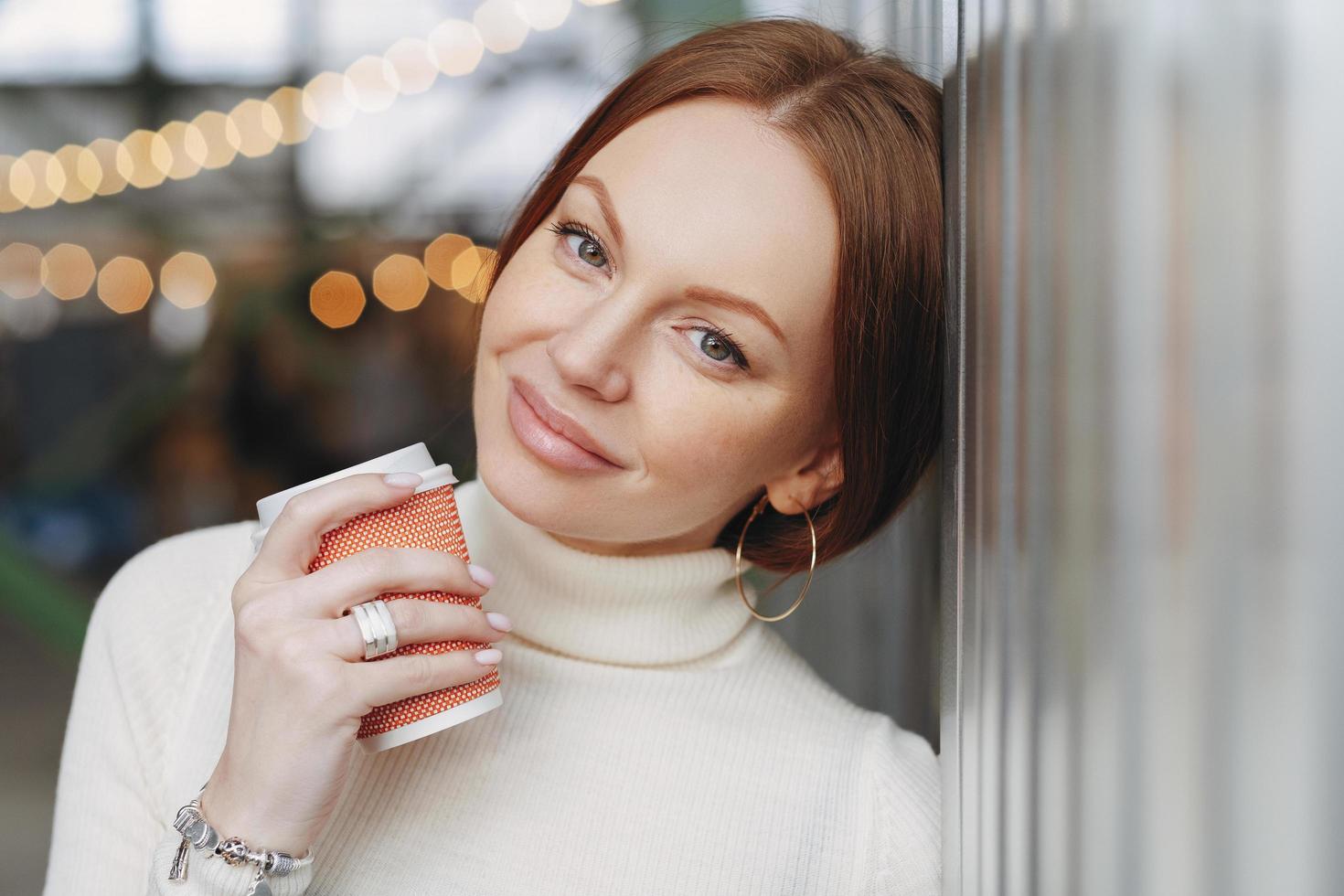 mujer caucásica soñadora y pensativa con maquillaje, vestida con ropa informal, se apoya en la pared, toma café para llevar, recuerda momentos agradables durante el paseo al aire libre, tiene un aspecto atractivo. concepto de bebida foto