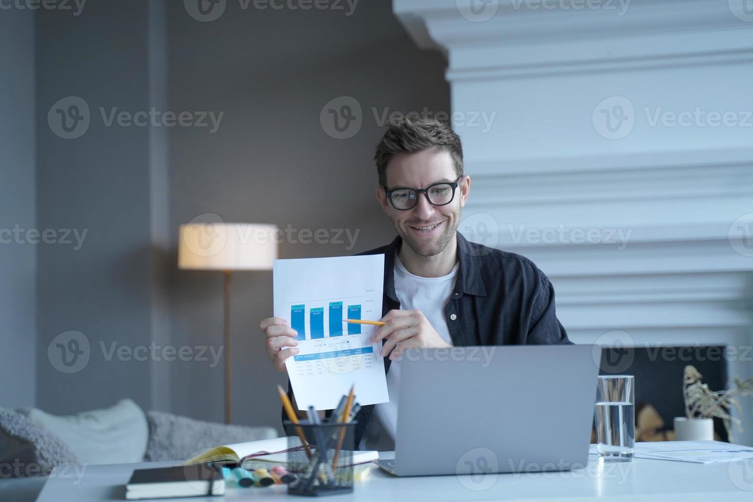 Young smiling german businessman pointing at document with graphics during internet meeting photo