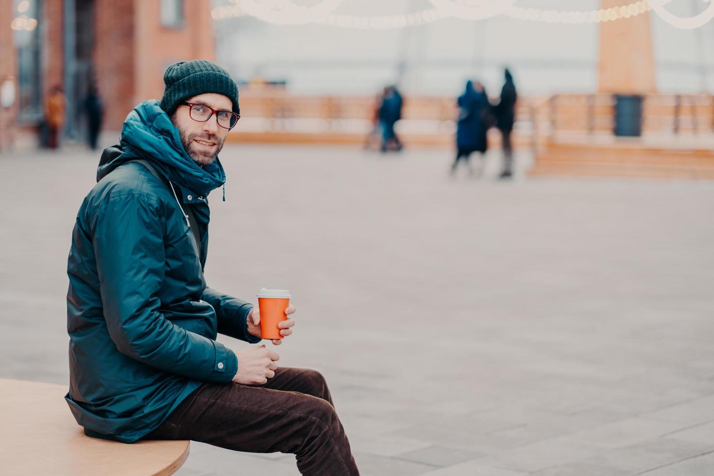 Sideways shot of handsome male adult dressed in street clothes, enjoys aromatic beverage, poses outdoor against blurred background with copy space for your promotion. People and rest concept photo