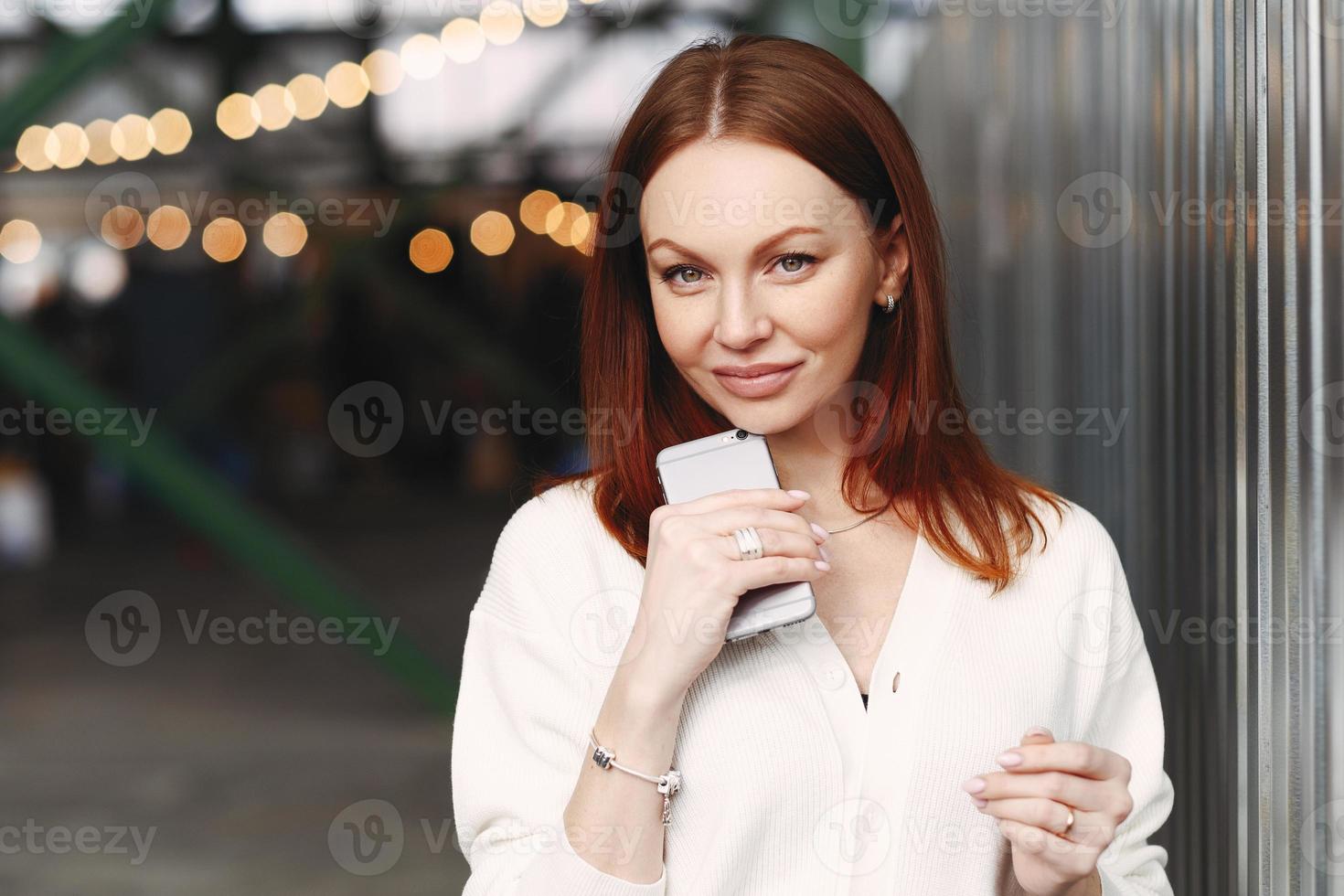 Photo of prosperous female business worker has break after work, sends text messages, holds cell phone in hand, dressed in white shirt, has browh hair, stands outdoor against blurred background