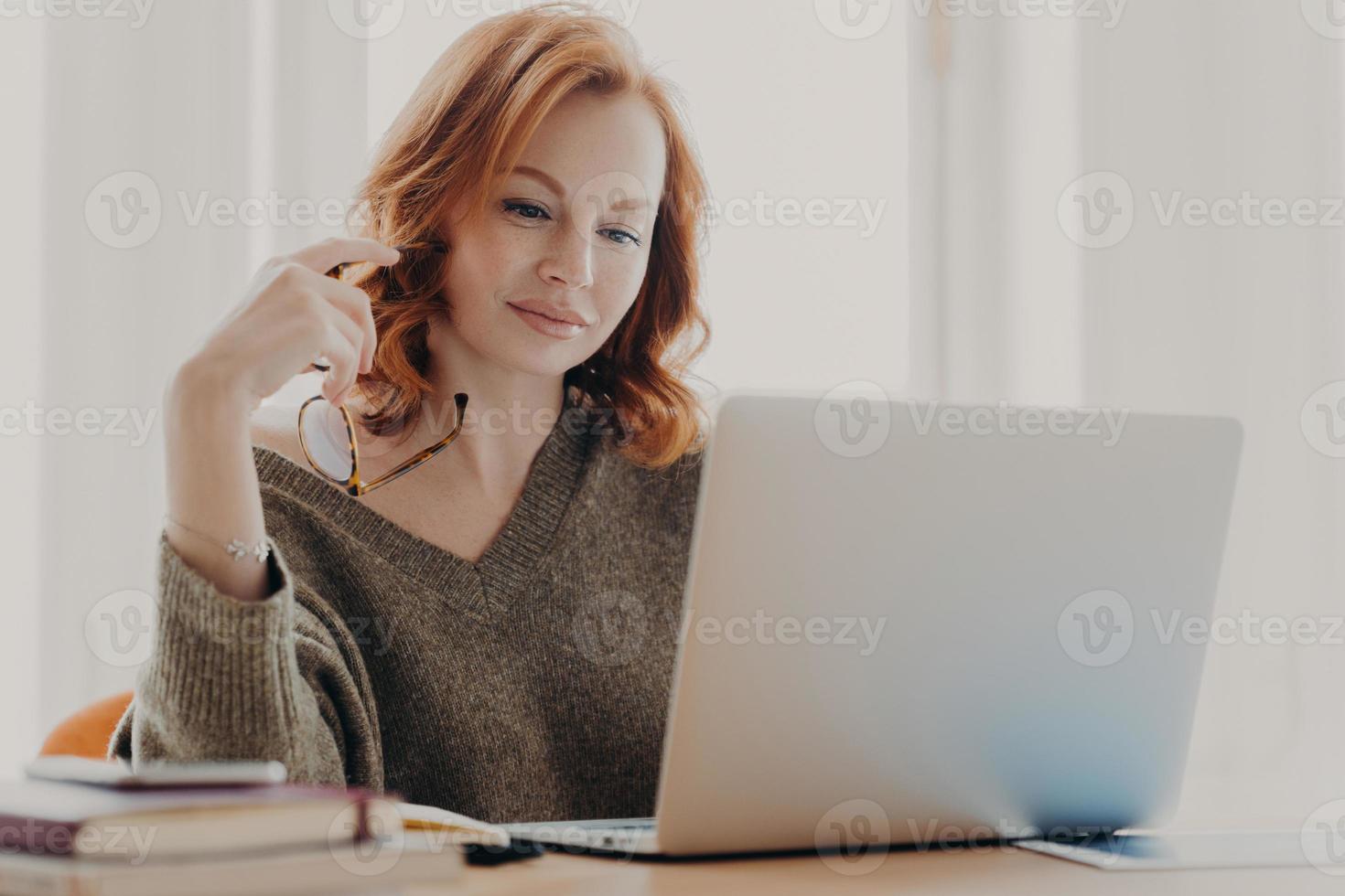 Concentrated female student reads information and checks data, focused at laptop screen, uses wireless internet connection, prepares for exam, poses at workplace, holds eyewear for vision correction photo