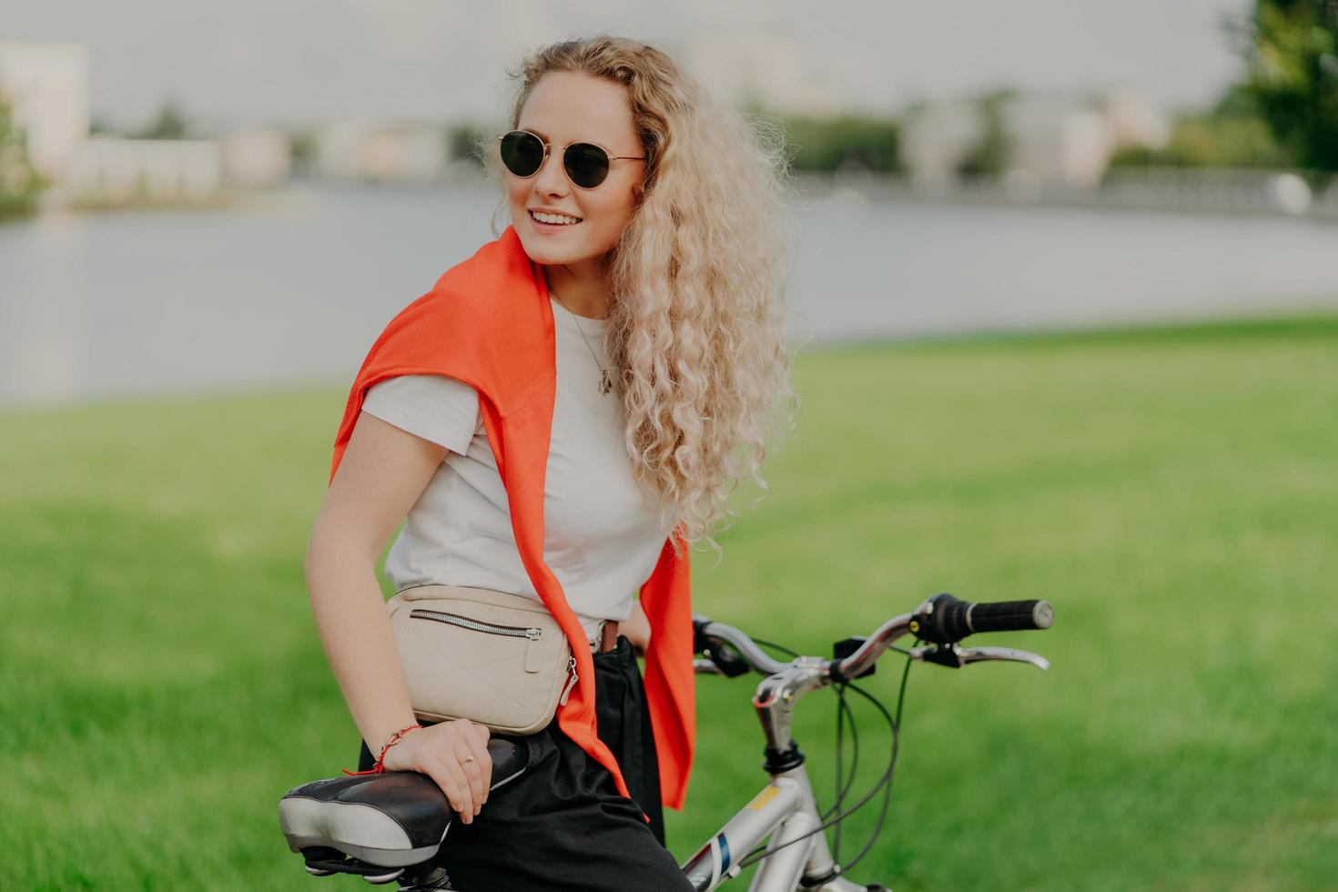 Active lifestyle concept. Positive young woman with curly light hair, rides bike on green lawn, wears shades, white t shirt and red sweater on shoulders, carries little bag, being in good mood photo