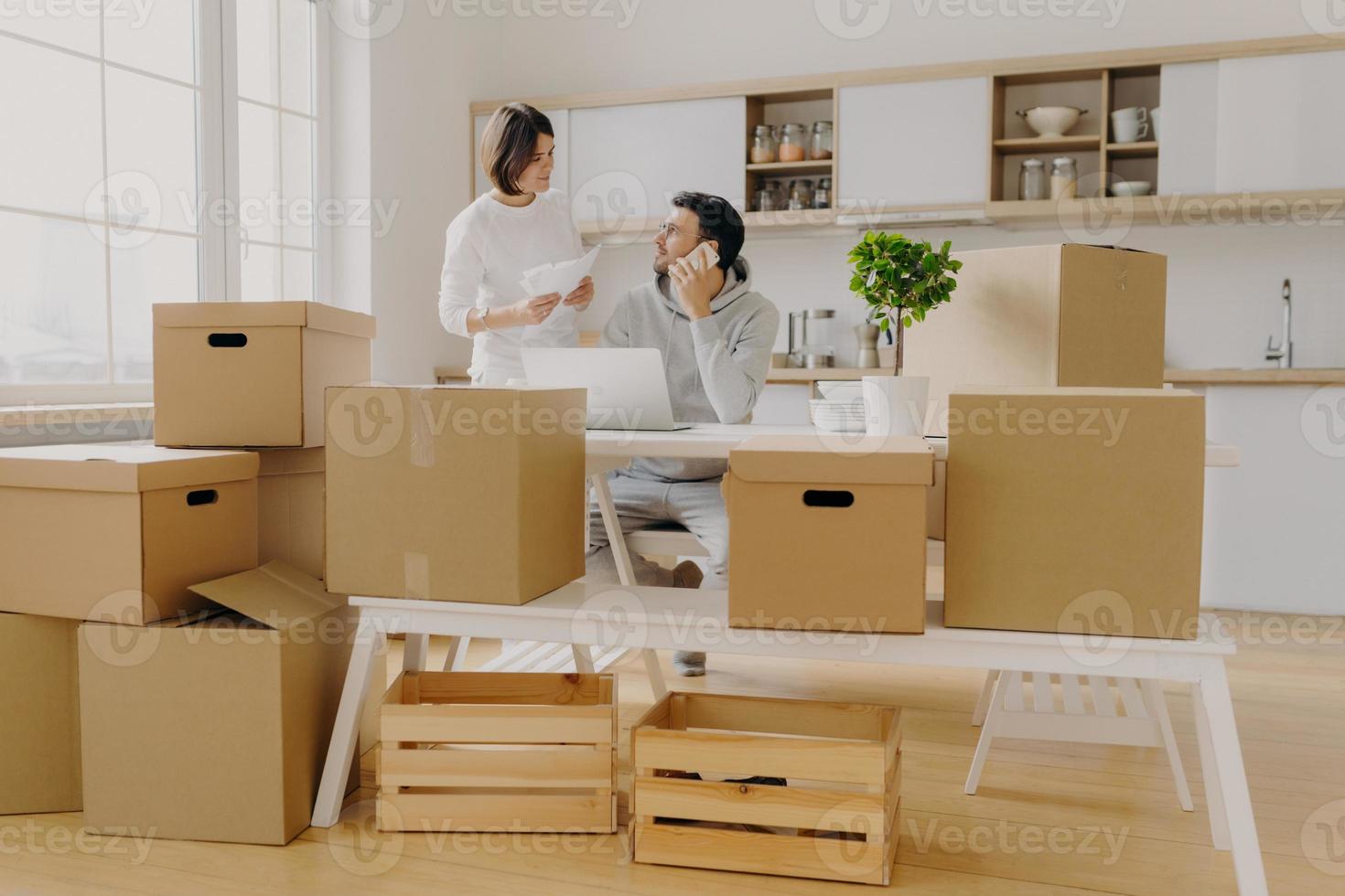 Family review their bank account, get loan application, talk about expenses, man talks via cellphone, sits at desk with laptop computer woman holds papers pose against kitchen interior with big window photo