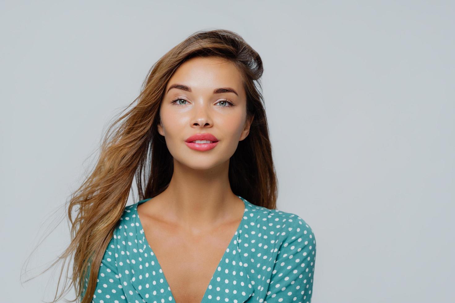 Portrait of pleasant looking young woman looks calmly at camera, has long hair, dressed in polka dot blouse, poses against white background, has natural beauty, has well cared face and makeup photo