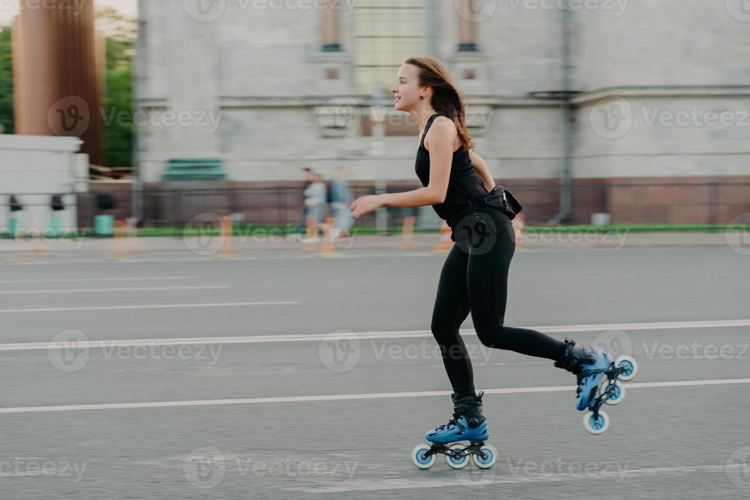 Summer rollerblading and fitness concept. Fit young dark haired woman fond of rollerskating exercises all body poses on road along asphalt during sunny day wears comfortable clothes has sporty figure photo