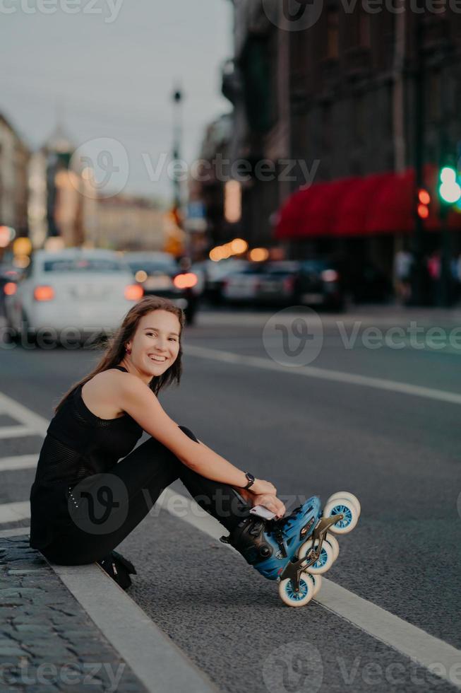 Outdoor shot of active slim woman poses on asphalt puts on rollerskates being in good mood spends free time riding rollerblades in urban place. Blurred city background. Hobby and recreation. photo