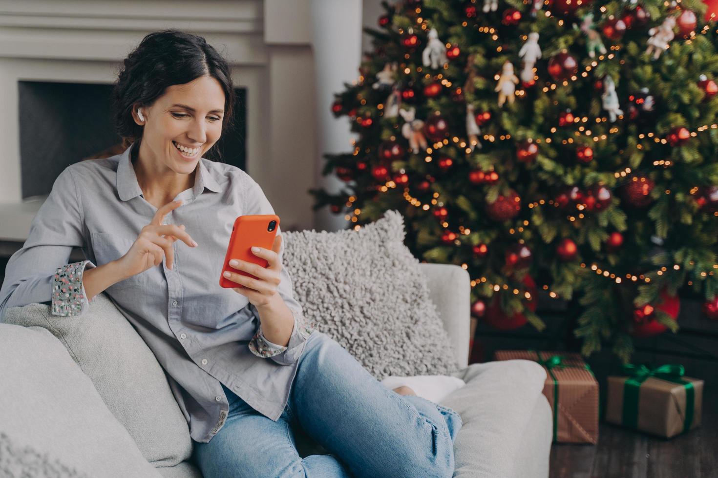 Happy female in earbuds holding smartphone, talking with family or friends online at Christmas time photo