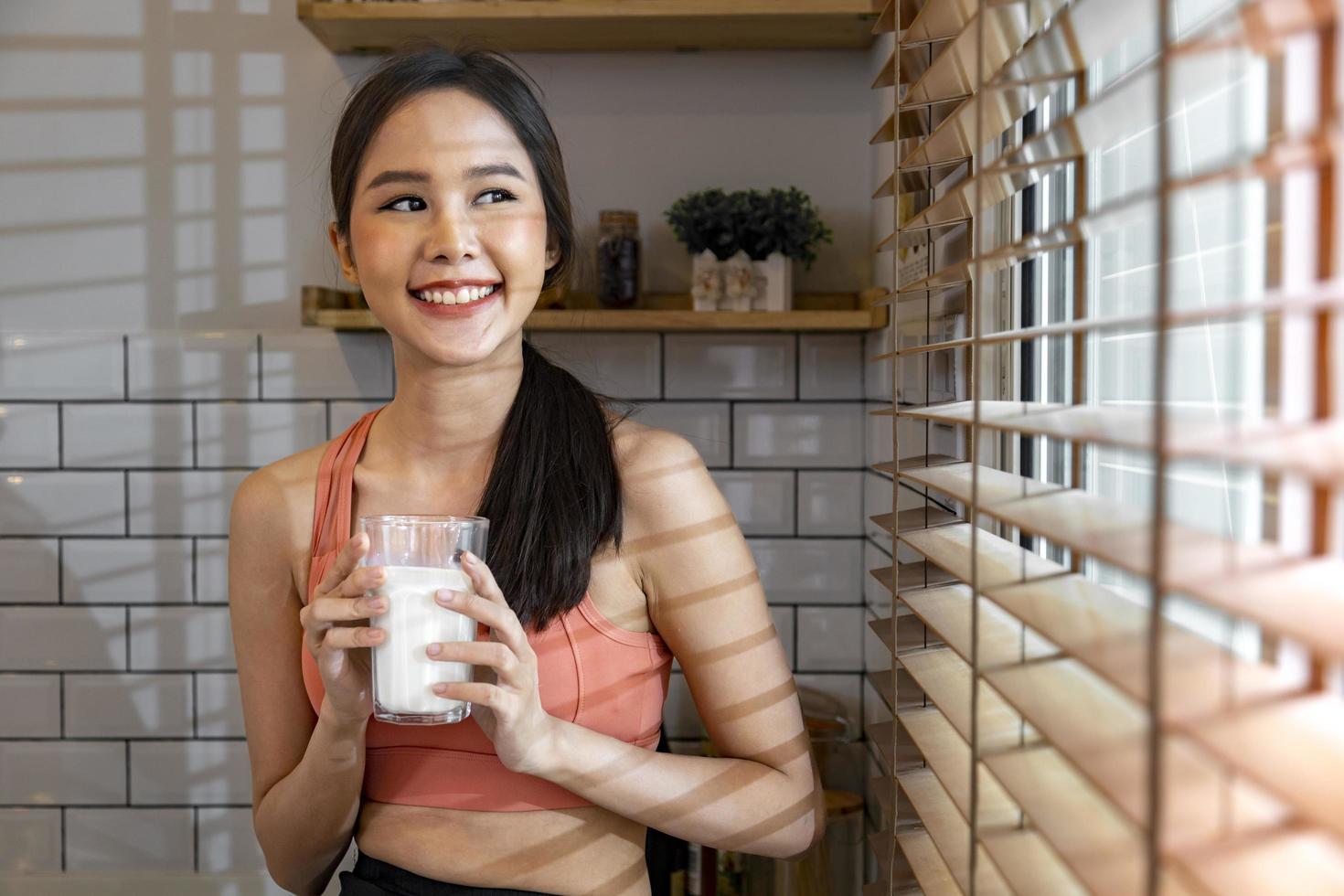 Healthy asian woman drinking a glass of dairy milk for while wearing sportswear for nutrition and protein supplement at the window for pregnancy and maternity benefit photo