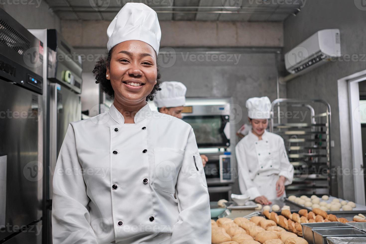 Portrait of young African American female chef in white cooking uniform looking at camera, cheerful smile with foods professional occupation, commercial pastry culinary jobs in a restaurant kitchen. photo