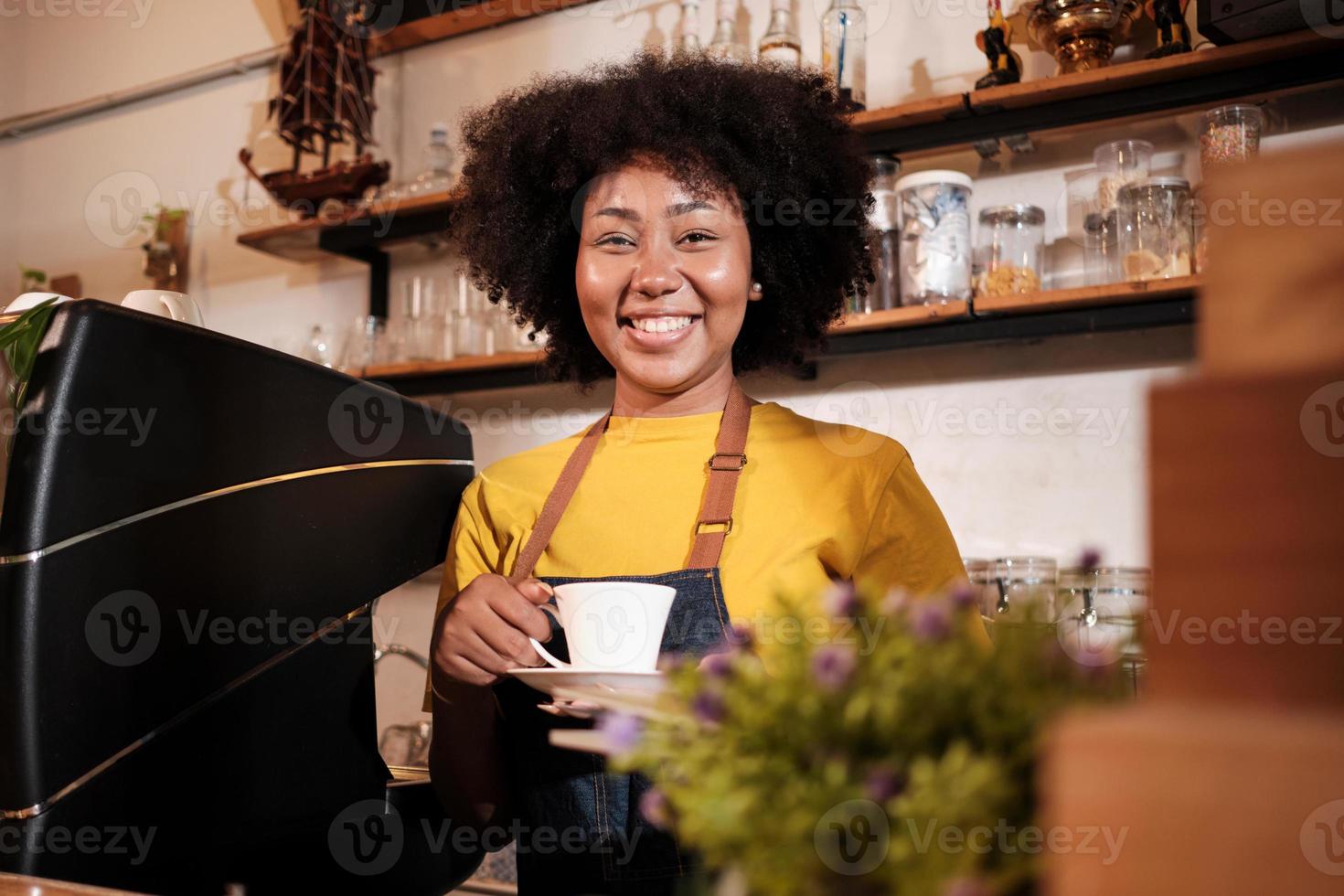 la barista afroamericana mira a la cámara, ofrece una taza de café al cliente con una sonrisa alegre, el servicio feliz trabaja en un restaurante informal, una joven emprendedora de pequeñas empresas. foto