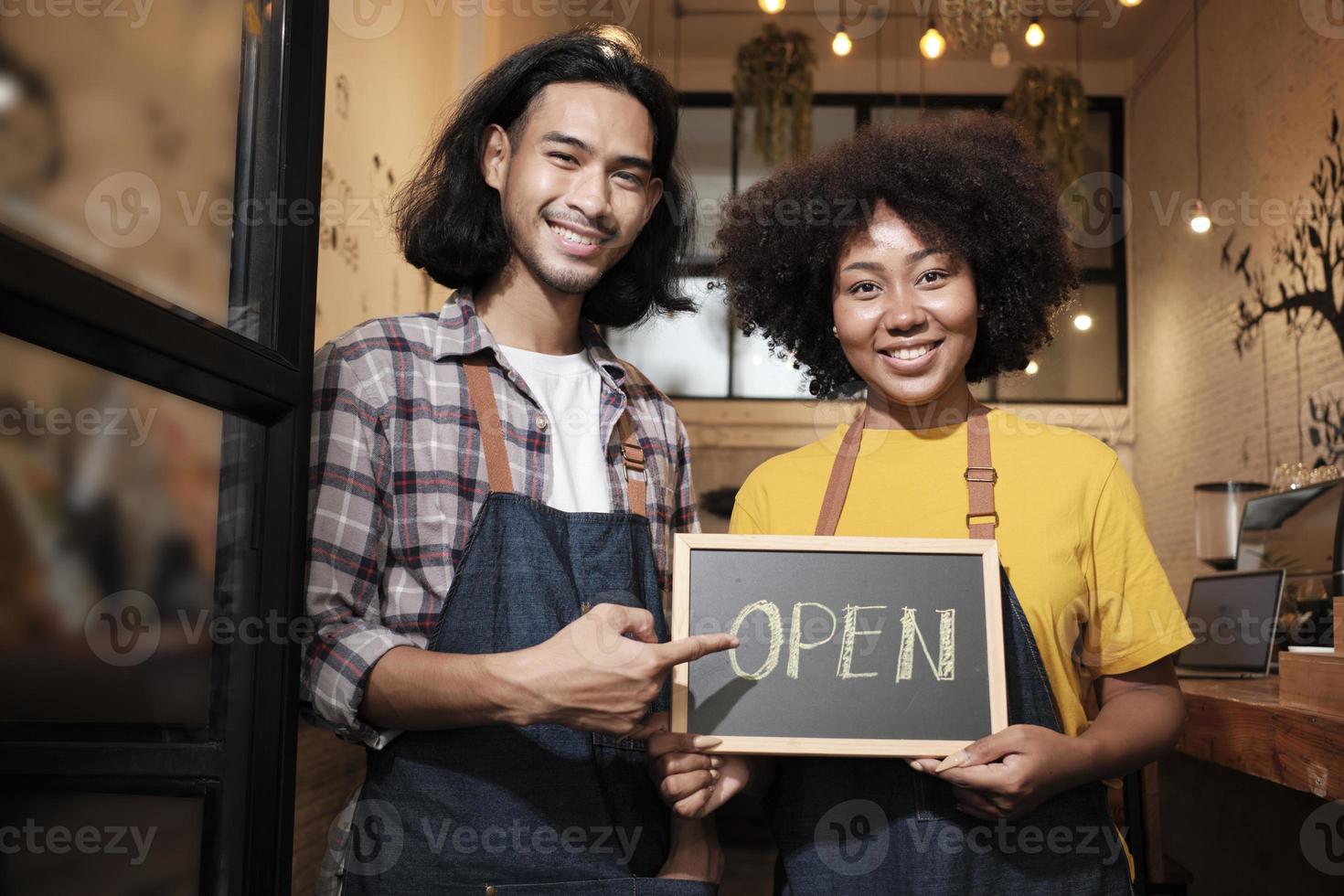 dos jóvenes socios baristas de inicio con delantales se paran en la puerta de un café informal, cartas a bordo y muestran un cartel abierto, sonrisas felices y alegres con trabajos de servicio de cafetería y nuevos empresarios. foto