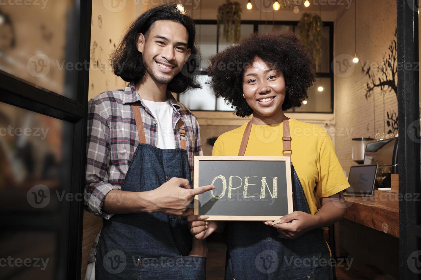 Two young startup barista partners with aprons stand at casual cafe door, letters on board and show open sign, happy and cheerful smiles with coffee shop service jobs, and new business entrepreneurs. photo