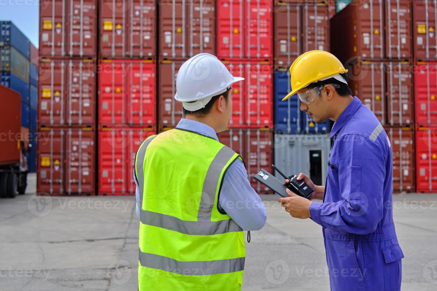 Two professional Asian male workers in safety uniforms and hard hats work at a logistics terminal with many stacks of containers, loading control shipping goods for the cargo transportation industry. photo