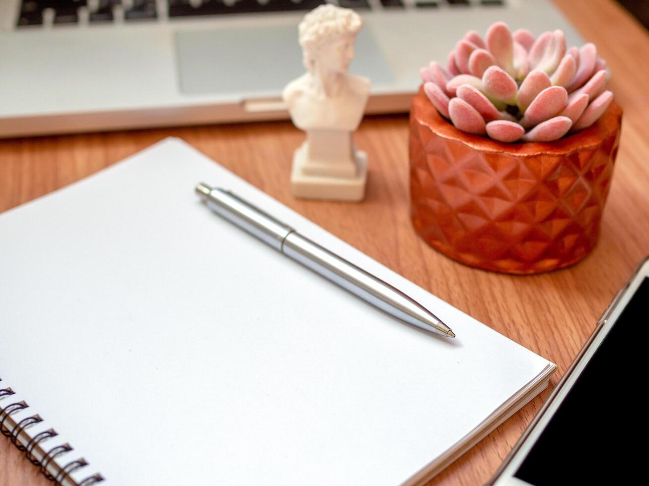 cuaderno en blanco con teléfono inteligente, computadora portátil y planta suculenta en una olla de cobre sobre fondo de mesa de madera foto