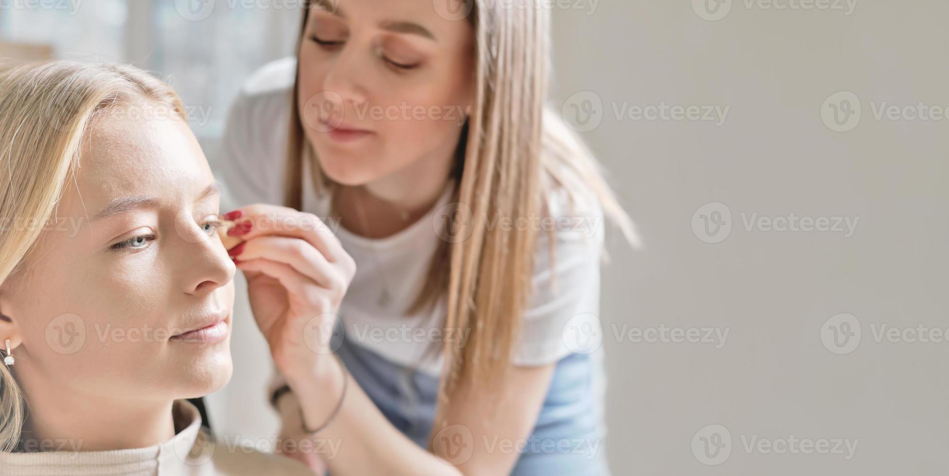 blond young woman model sitting in a beauty studio. make-up artist applying CC cream on her face. skin correction. make up in salon.copy space photo