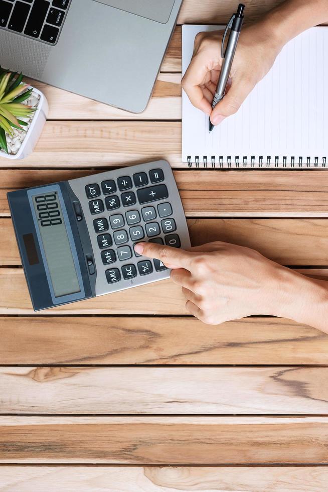 Top view Businesswoman hand writing and calculating with calculator, pen, computer laptop and plant pot on wood office desk table background. workspace or home office with copy space for text concept photo