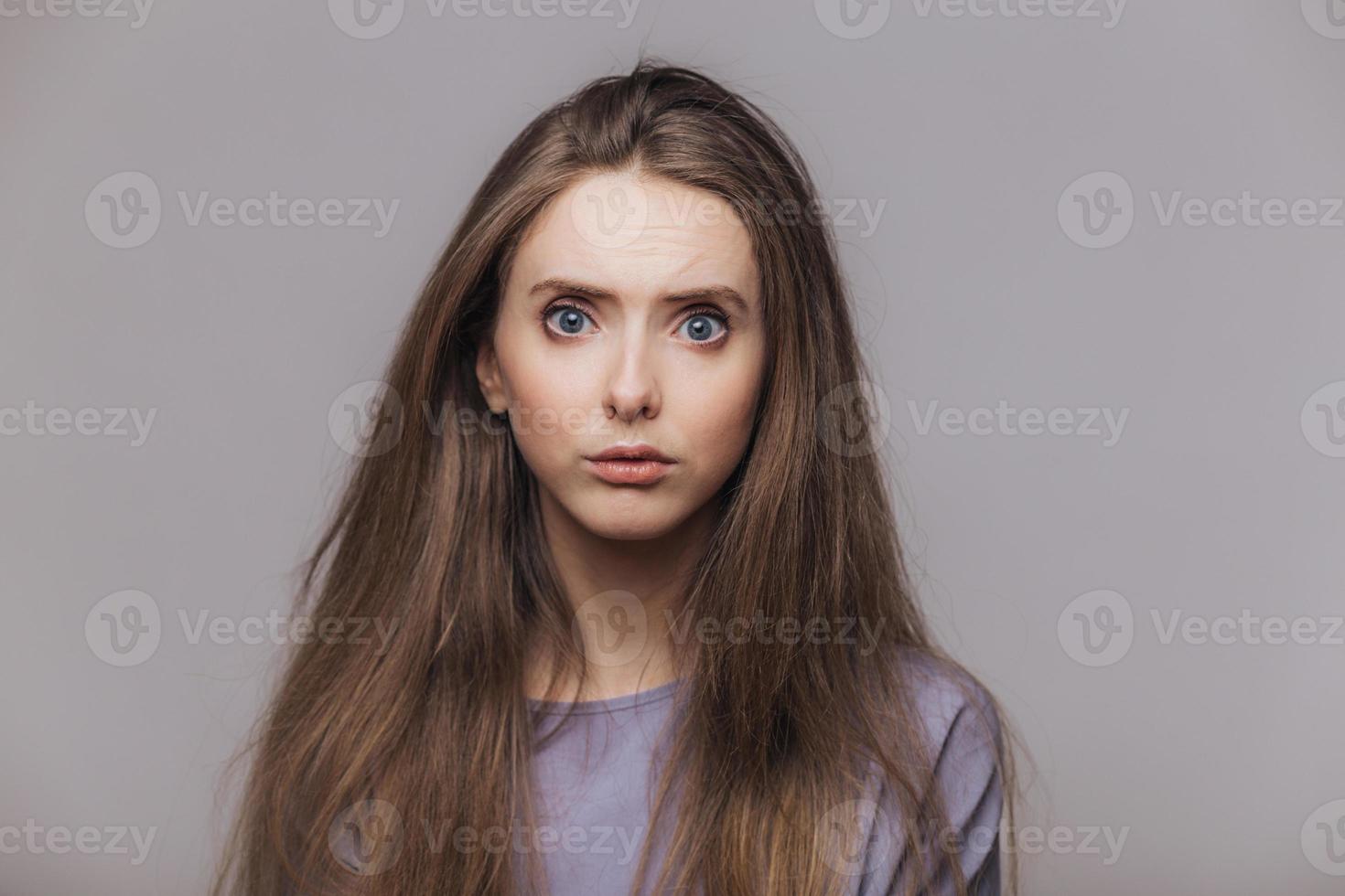 foto de estudio de una modelo femenina de ojos azules de aspecto agradable con cabello largo oscuro, tiene una expresión desconcertada al escuchar noticias impactantes de un interlocutor, aislado sobre fondo gris. concepto de expresiones faciales