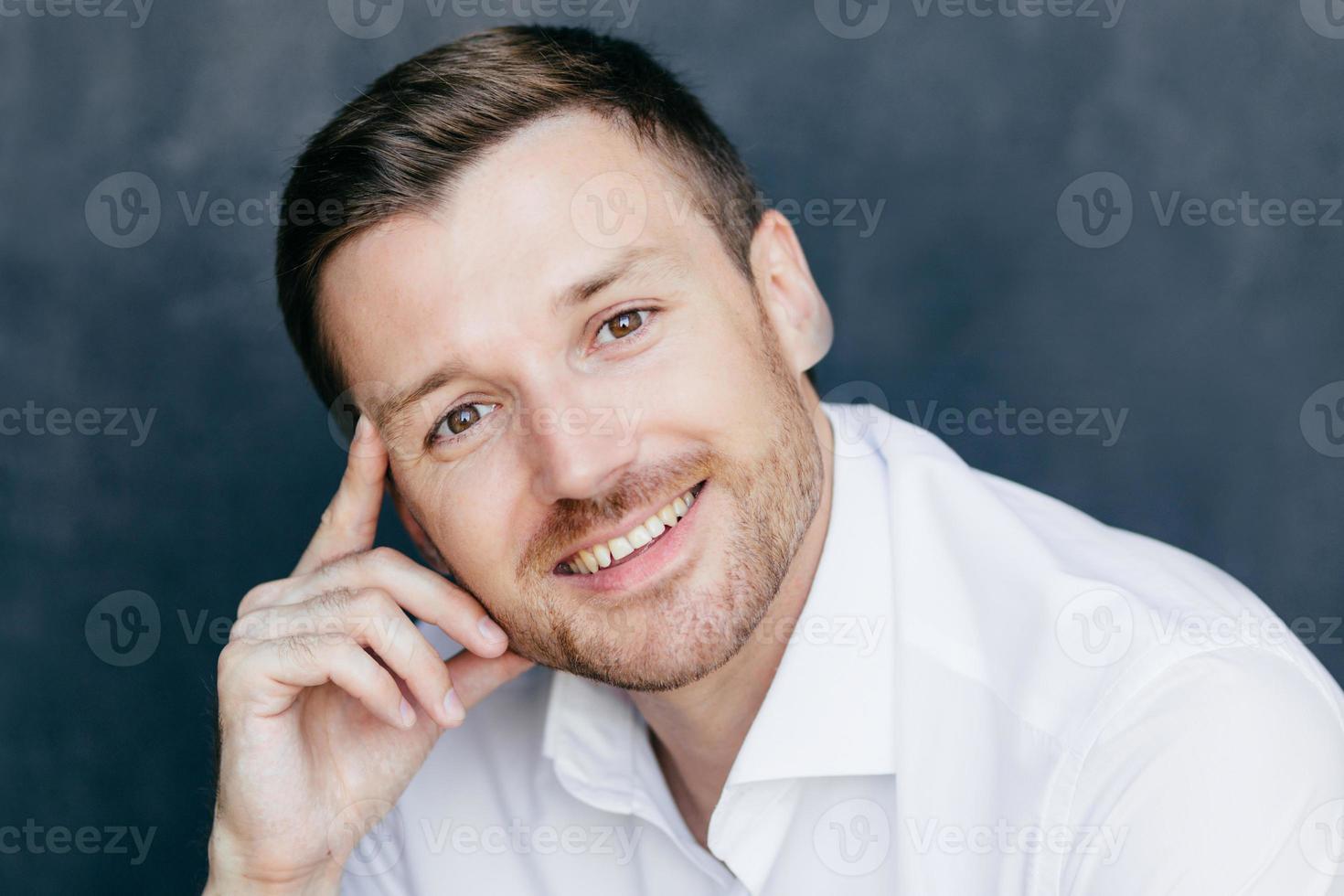 Cheerful male employee has joyful expression, keeps finger on temple, rejoices starting vacation, dressed in formal white shirt, poses against dark background. Successful businessman indoor. photo