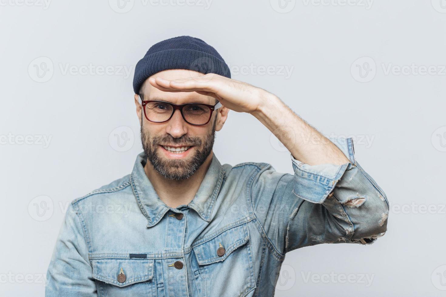 Cheerful unshaven middle aged male with thick stubble looks happily into distance, keeps hand near orehead, tries to see something, wears eyewear and denim jacket, isolated over grey background. photo