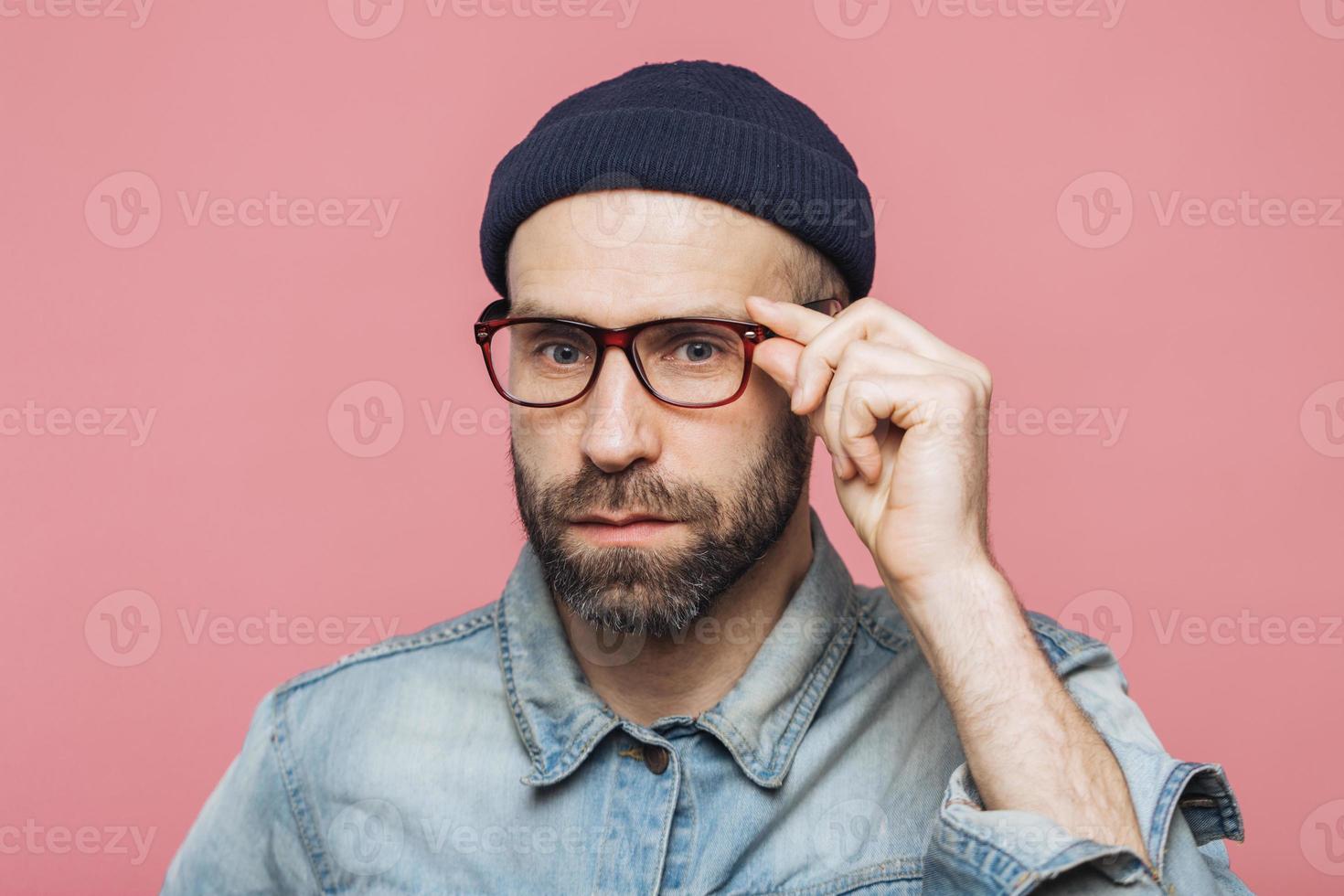 un apuesto hombre barbudo con expresión seria usa anteojos y anteojos, vestido con una camisa de mezclilla a la moda, aislado sobre fondo rosa. concepto de personas, expresiones faciales y emociones. foto