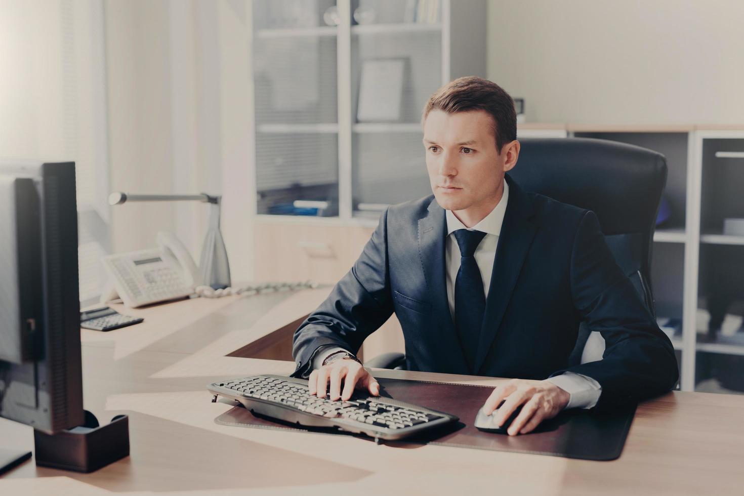 Serious concentrated businessman sits in front of computer at work place, keyboards information, wears suit, prepares business project for next year, poses in spacious cabinet. People and finances photo