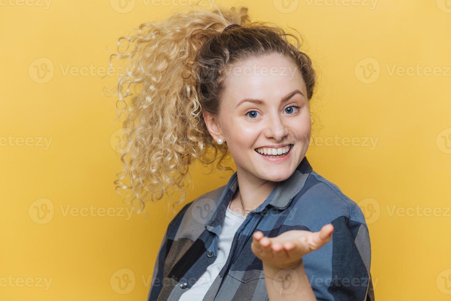 encantadora mujer alegre estira la mano en primer plano, tiene una amplia sonrisa, demuestra dientes blancos perfectos, usa camisa a cuadros, posa contra un fondo amarillo. sonriente satisfecha modelo femenino interior foto