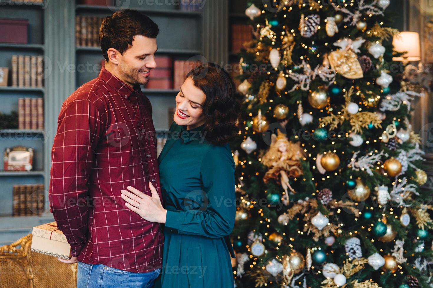Lovely family couple pretty wife stands near her husband who prepares New Year present for her. Good relationnship concept. Woman and man stand against decorated Christmas tree. Winter holidays photo